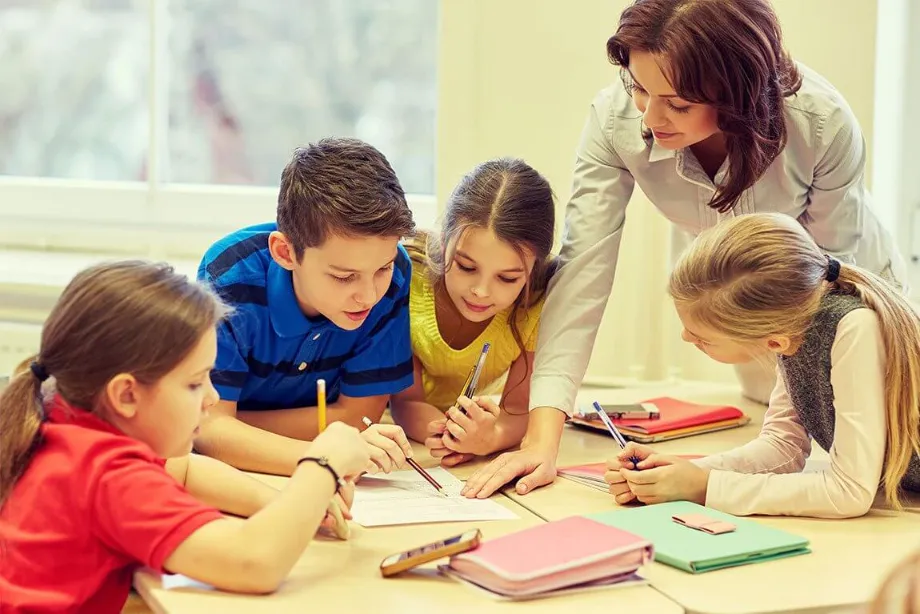 Lehrerin hilft vier Grundschulkindern beim gemeinsamen Schreiben am Tisch